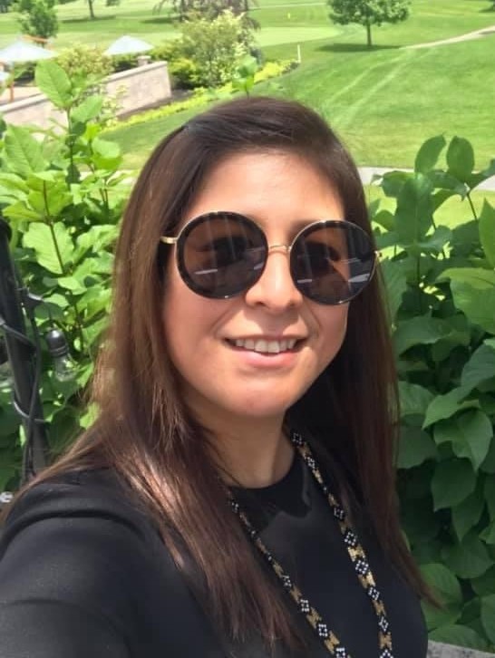 Image of a woman with brown hair, wearing sunglases and a dark top, facing the camera and smiling