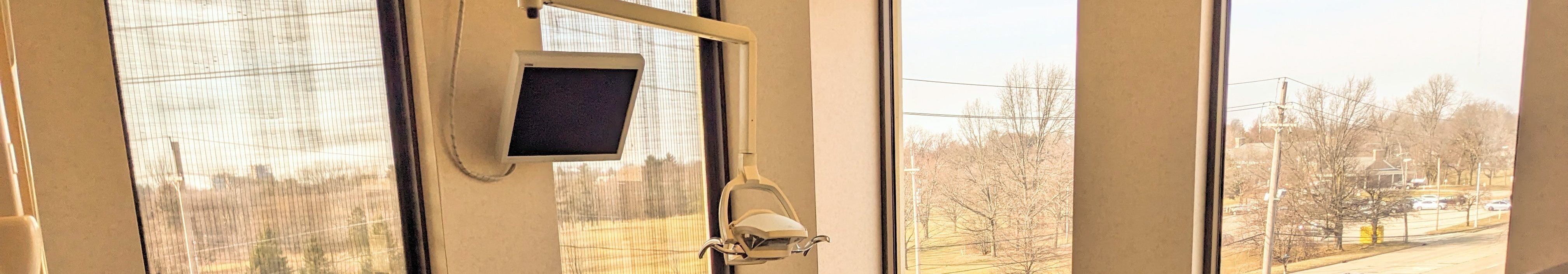 Snippet of an image of a dental operatory, there is a ceiling-mounted computer monitor on the upper right, and four windows that look out on treetops