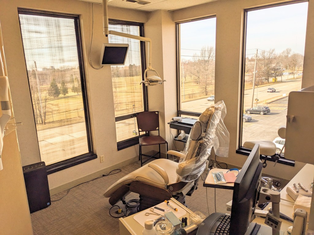 Snippet of an image of a dental operatory, there is a patient chair in the center surrounded by windows that look out on a park on the left and a parking lot on the right